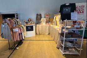 Vendor setup with clothing and merchandise on tables and racks in a room.